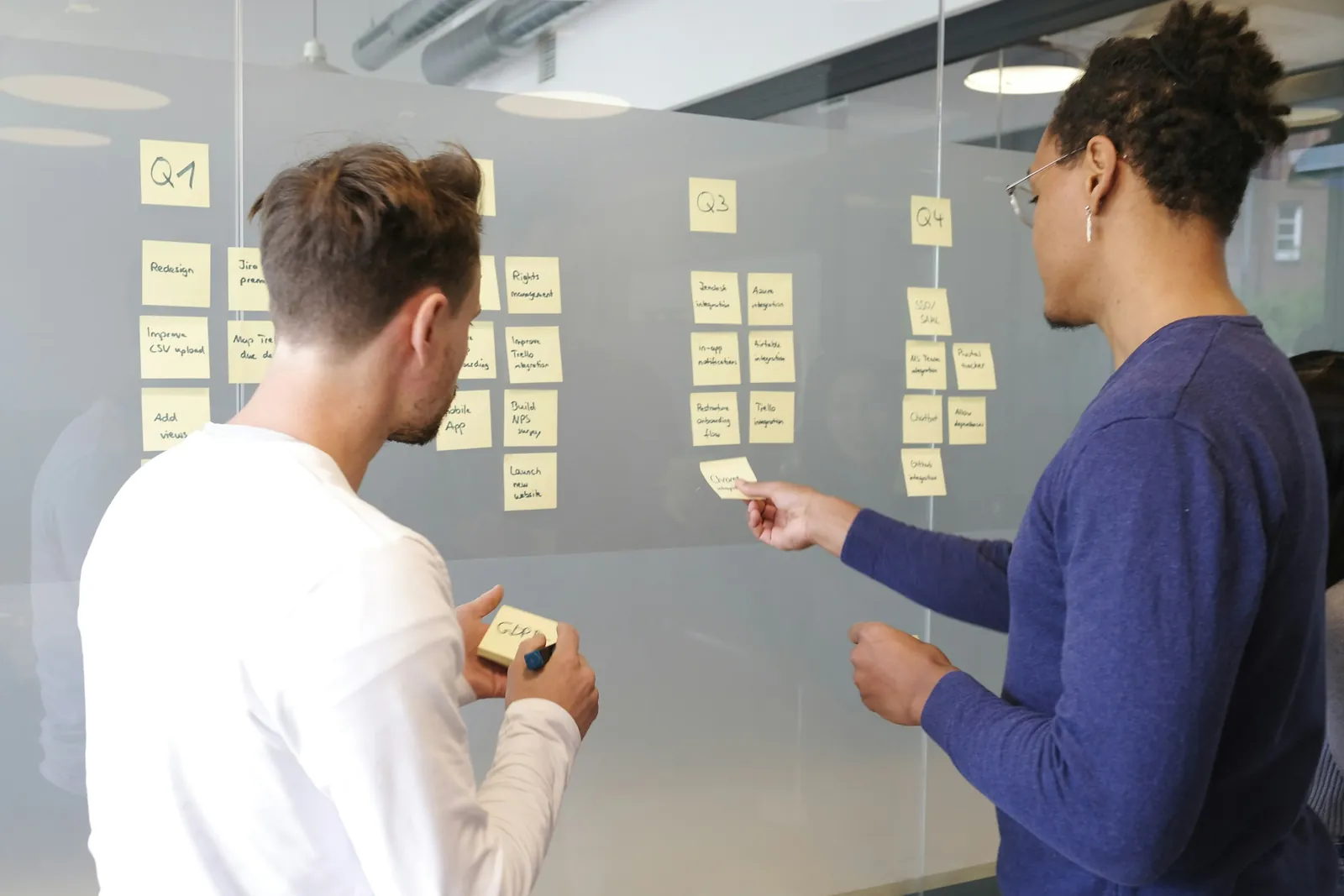 Two colleagues arranging handwritten sticky notes into columns on a glass planning wall