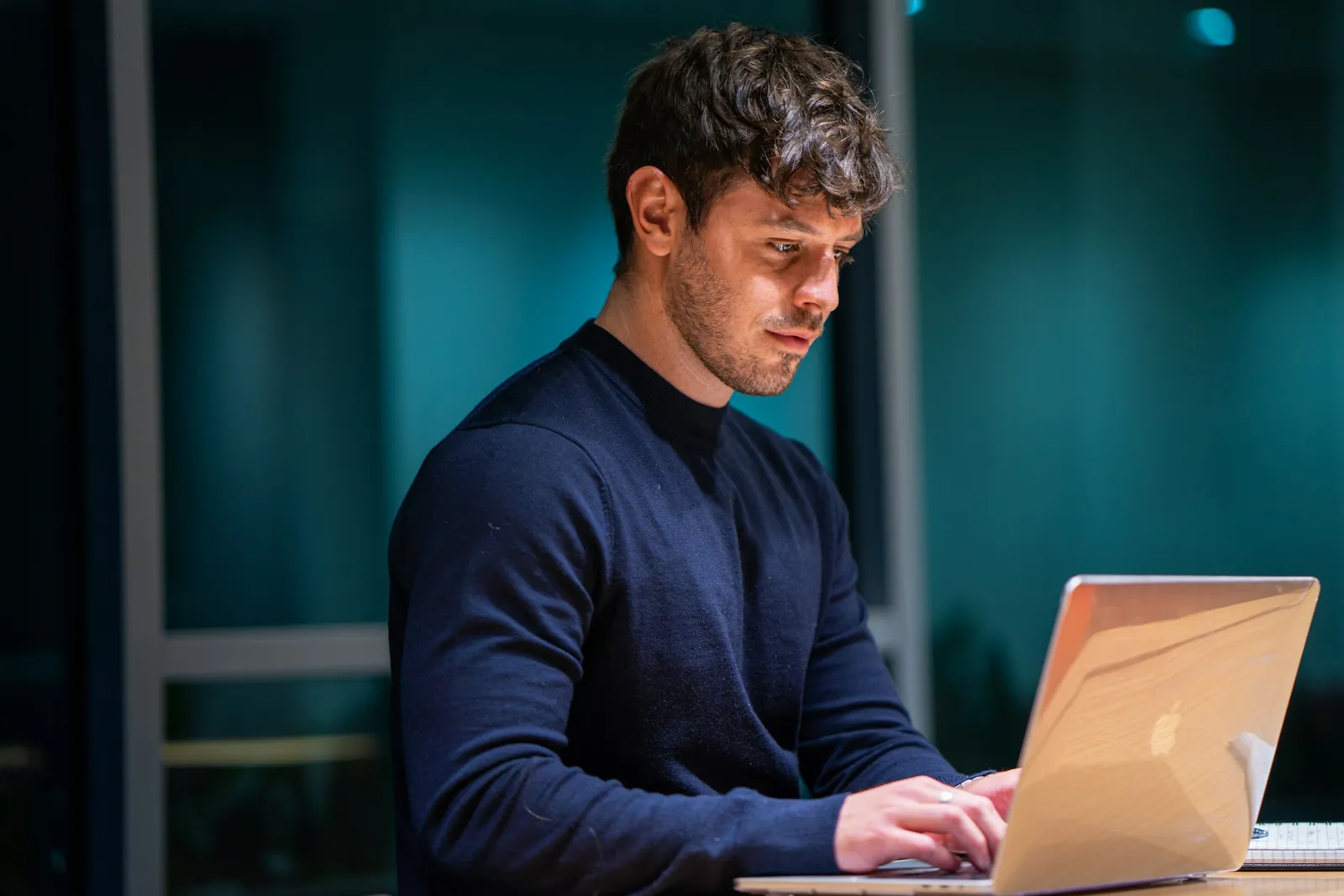 A man in a navy sweater reading carefully at a laptop in a dim office