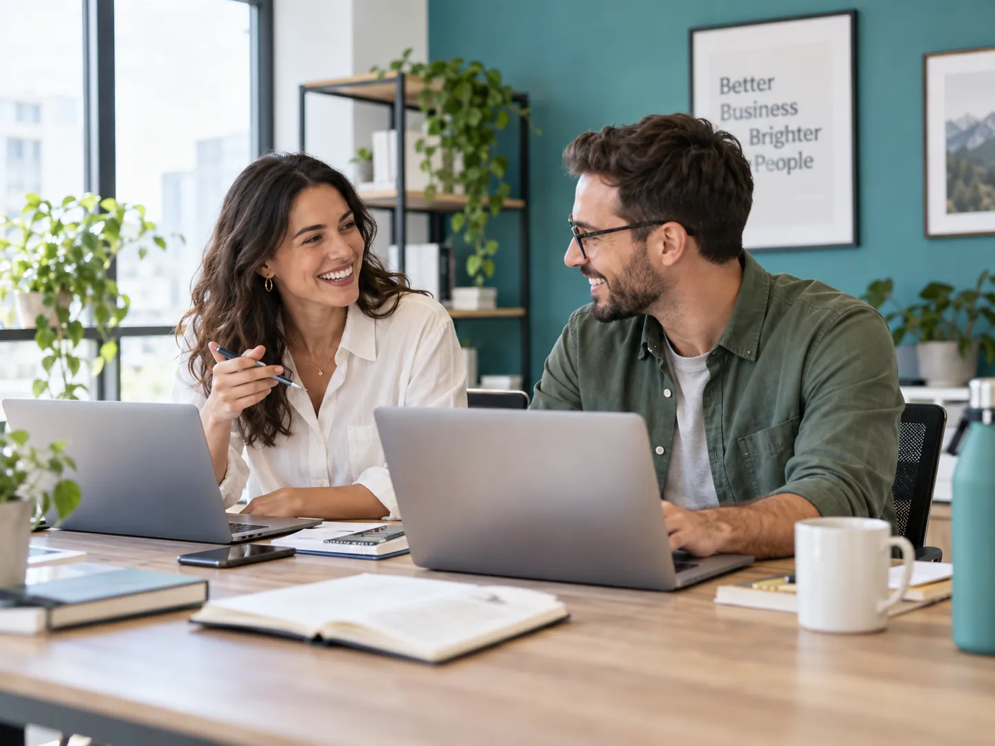 Two colleagues smiling as they work side by side on laptops in a sunny office